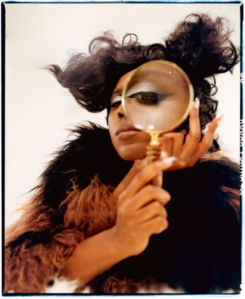 portrait of comedian ziwe holding a magnifying glass up to one eye. she wears a brown and black fur coat and winged liner.