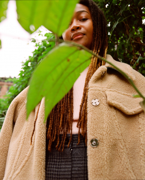 portrait of a person with long ombre locs partially obscured through a green leaf. they wear a tan fleece coat.