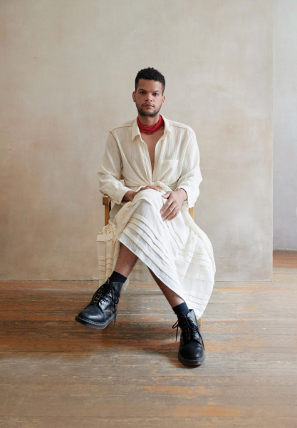 an afro mexican person sitting on a stool with legs crossed wearing a cream skirt and matching button down with black...